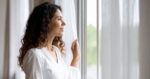 A woman with curly hair stands indoors, looking out a window while holding a sheer curtain with one hand.