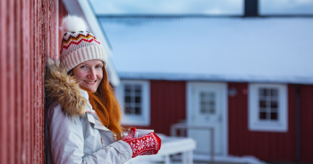 A woman in winter clothing stands outside a red building with snow on the roof, holding a mug and smiling at the camera.