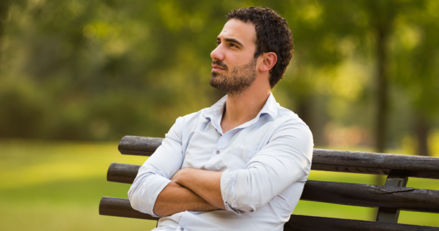 A man with dark hair and a beard sits on a bench outdoors with his arms crossed, looking thoughtfully into the distance. Trees and greenery are visible in the background.