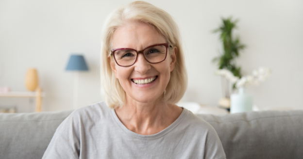 Older woman with glasses and gray shirt smiling while sitting on a couch in a bright living room with plants and decor in the background.