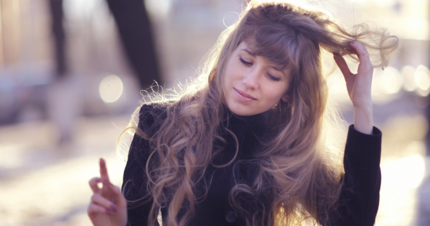 Woman with long wavy hair wearing a black coat stands outdoors, eyes closed, holding up her hair with one hand and raising the other finger, with a blurred background.