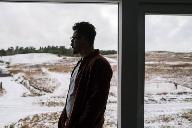 A man sits alone looking out a window at a snowy landscape, a representative image, not a photograph of any person discussed in the article.