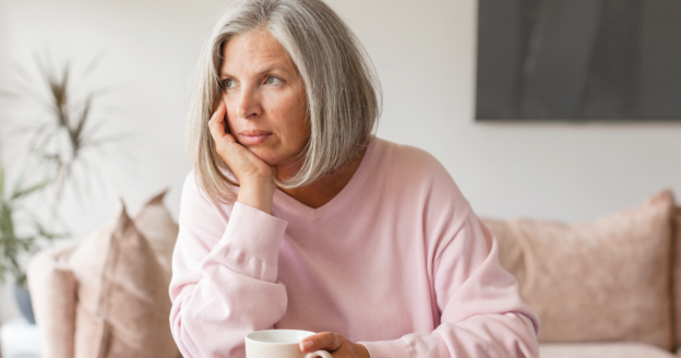 Woman with gray hair in a pink sweater sits indoors, resting her chin on her hand and holding a white mug, looking thoughtfully into the distance.