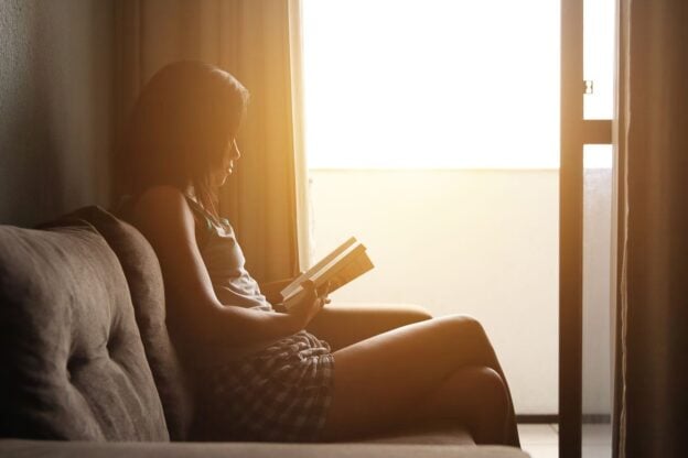 A woman reading alone on a sofa beside a sunlit window