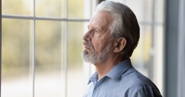 Older man with gray hair and beard looks thoughtfully out a window. He wears a blue collared shirt and stands in a softly lit room.