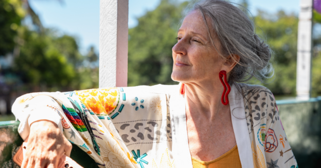 Older woman with gray hair in a bun, wearing a patterned cardigan and red earrings, sits outdoors in sunlight, looking thoughtfully into the distance. Green trees are in the background.