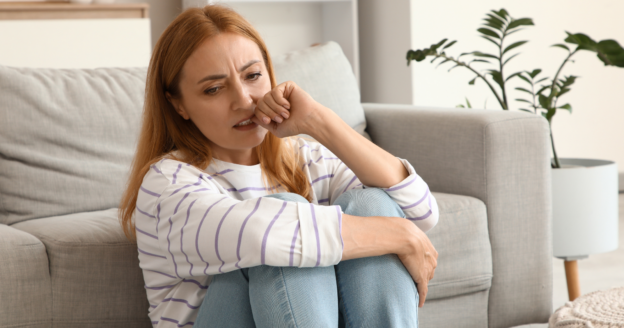 Woman with red hair sits on the floor by a sofa, hugging her knees and looking pensive while biting her hand. A green plant and neutral decor are in the background.