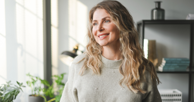 A woman with wavy blonde hair in a light sweater stands indoors near a window, smiling. There are plants and shelves with books in the background.
