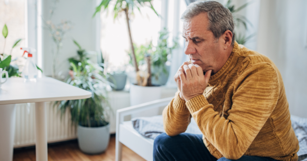 An older man in a mustard sweater sits on a bed, leaning forward with hands clasped, appearing deep in thought in a bright room with plants.