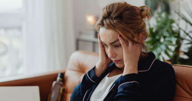A woman sits on a couch with her hands on her temples, appearing stressed or deep in thought.