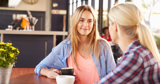 Two women sit at a table in a café, having a conversation. One woman holds a cup, and there are cups and a small plant on the table. Natural light comes through large windows.