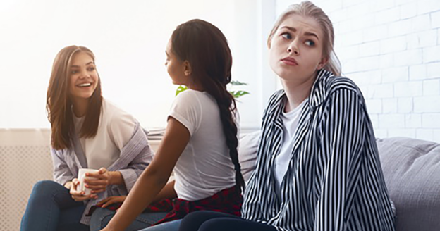 Three young women sit on a couch; two are talking and smiling, while the third woman looks away with a displeased or excluded expression.
