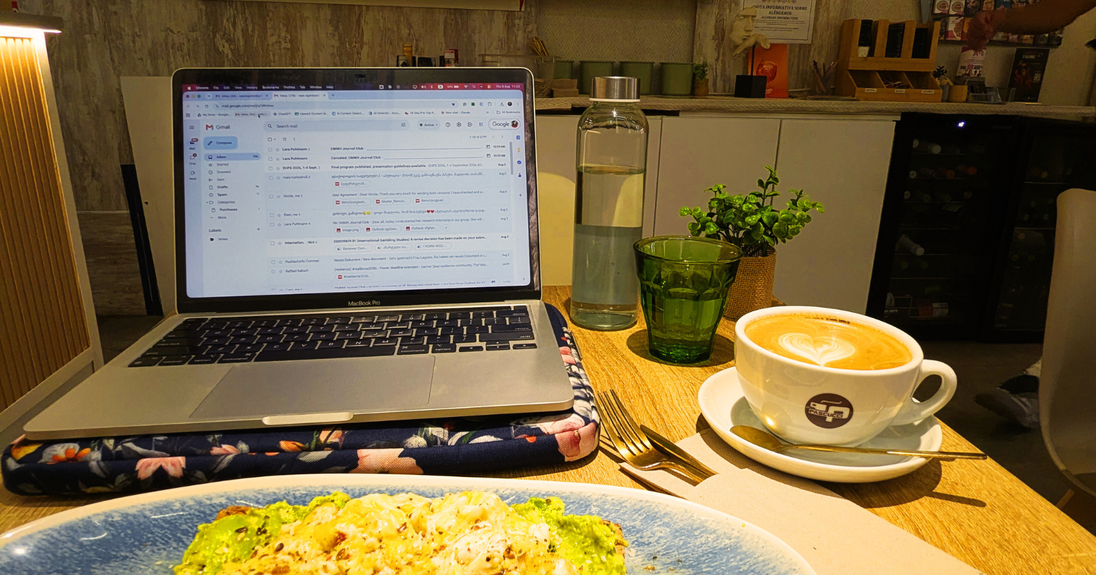 A laptop displaying an email inbox sits on a wooden table with a cappuccino, a green glass, a water bottle, a potted plant, and a plate of avocado toast in a café setting.