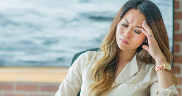 A woman with long hair sits indoors, eyes closed and hand to her forehead, appearing to have a headache or to be stressed.