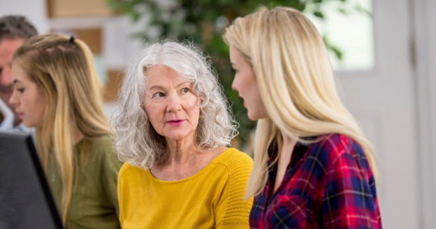 An older woman with gray hair talks to a younger blonde woman in a plaid shirt. Both are seated indoors, with other people and a computer monitor visible in the background.
