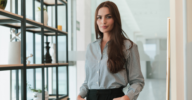 A woman with long brown hair stands in an office, wearing a striped shirt and black pants, with shelves and glass walls in the background.